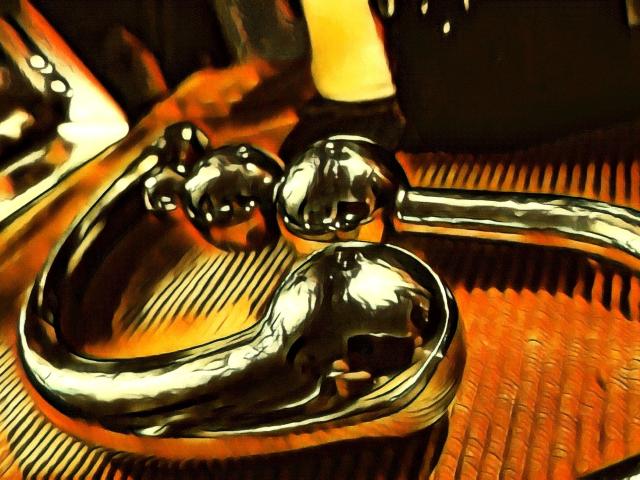 Shiny, metallic butt plug with two rounded, reflective knobs and a curved, ring-like handle. It's shiny and reflects light, resting on a textured, orange surface.