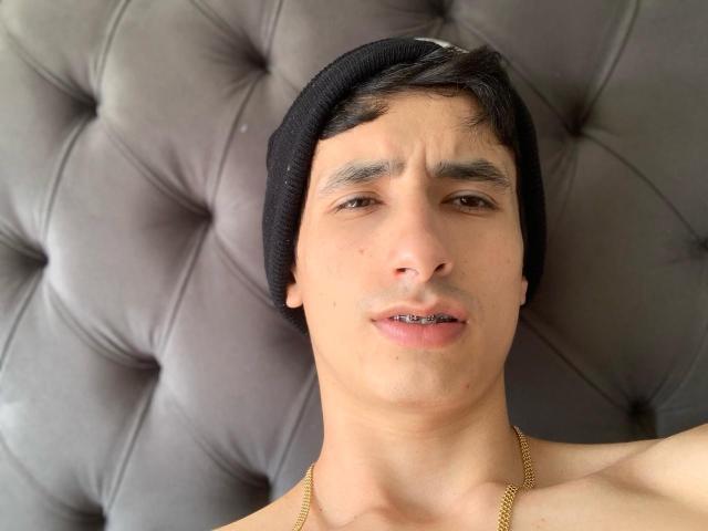 AlexArngo with a black beanie, brown eyes, and slight frown, shirtless, wearing a gold chain, lying on a tufted gray cushion.