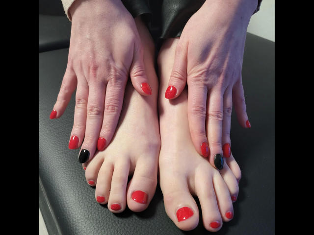 Zara-Bizarr's hands with red nail polish and one black nail, resting on her feet with matching red nail polish. Her feet are on a black leather surface.