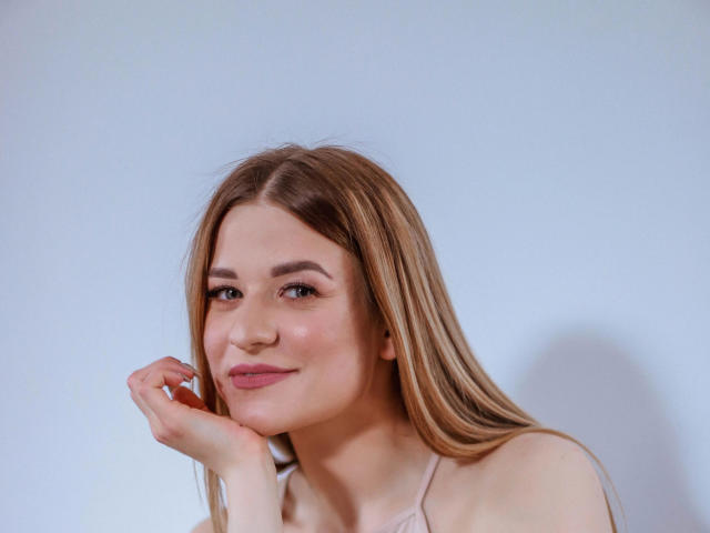 SourrSanndy-hot with long blonde hair, light skin, pink lips, subtle makeup, and a cute smile, resting her chin on her hand, wearing a pink top, in a portrait.