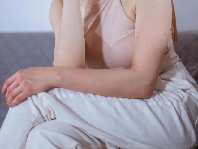 SourrSanndy-hot in a pink tank top, pale skin, blonde hair, sitting on a gray bed with white pants, hands resting on white bedding, soft light, relaxed.