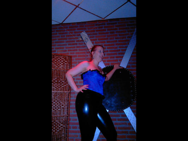 MistressOnline-hot with a blue corset, black shiny pants, and a shield, holding a sword, standing with her hand on her hip, looking confident. She's got a tattoo on her head.