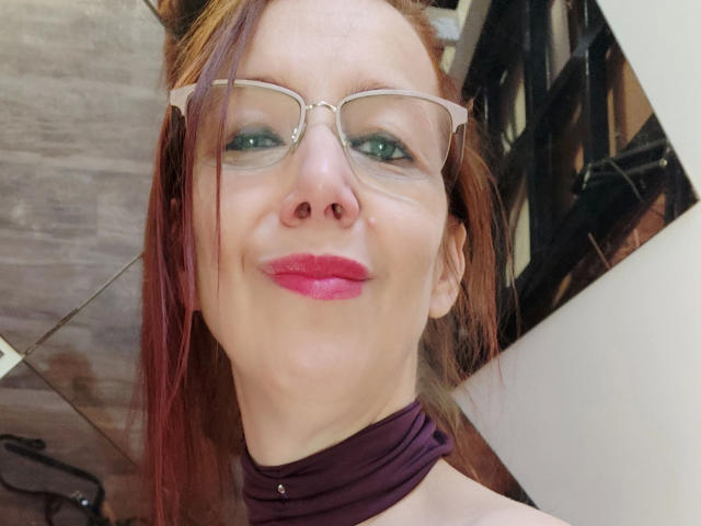 MistressOnline - sensual, usando óculos, batom rosa e um lenço roxo, sorrindo para a câmera. Ela tem cabelo ruivo, olhos verdes e um tom de pele claro..