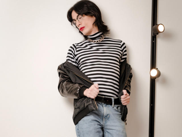 MilarHole with black and white striped turtleneck, jeans, and black leather jacket, pulling jacket off, wearing glasses, red lipstick, and chunky necklace, standing next to lit wall.
