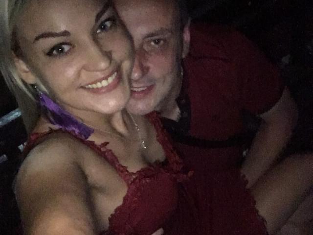 EvaAndAnto has blonde hair, purple earrings, and a red lace dress, smiling with her tits visible. Anto, with a red shirt, is behind her, looking happy.