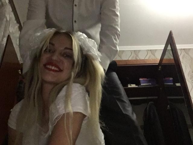 EvaAndAnto with long blonde hair, white dress, and flower headpiece, smiling, has her hair being held up by a guy in a white shirt, standing behind her.