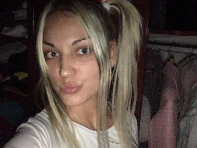 Blonde EvaAndAnto with puffy lips, white tee, and ponytails, looking at the camera with a slight smirk. Her tits are small and her pussy isn't visible.