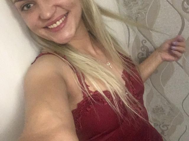 EvaAndAnto with blonde hair, smiling, wearing a red lace camisole, showing her tits, holding her hair, wearing a necklace, in a selfie, with a white wall.