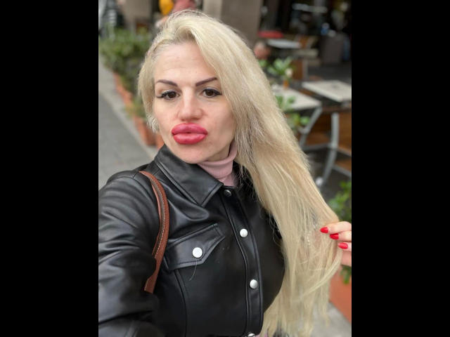 PamelaDeluxe-hot with long blonde hair, black leather jacket, pink scarf, and red lipstick, pouting her full pink lips, holding her hair with red nails.