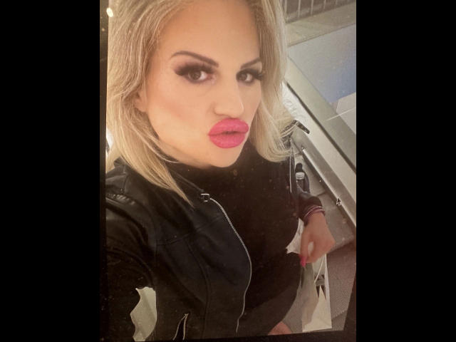 PamelaDeluxe-hot with blonde hair, pink lipstick, and heavy eyeliner, wearing a black jacket, holding shopping bags, sitting in a car, looking at the camera with a pouty face.