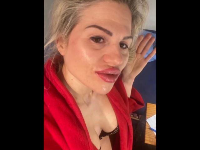 PamelaDeluxe-hot with blonde hair, red robe, wet skin, full lips, and visible tits, hand up, slightly smiling, looking at camera.