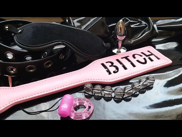 Pink dildo with "BITCH" on it, black leather belt, pink butt plug, and glass bead bracelet on shiny black and white surface. Circea-hot's sex toys.