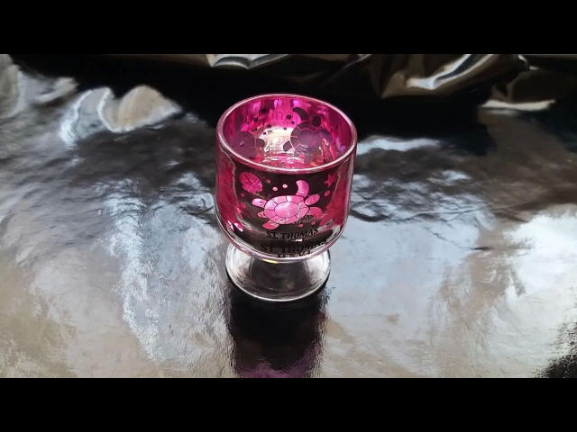 Un elegante candelabro de cristal rosa con un diseño floral, que se encuentra sobre una superficie negra reflectante. En su interior, tiene una pequeña vela blanca..