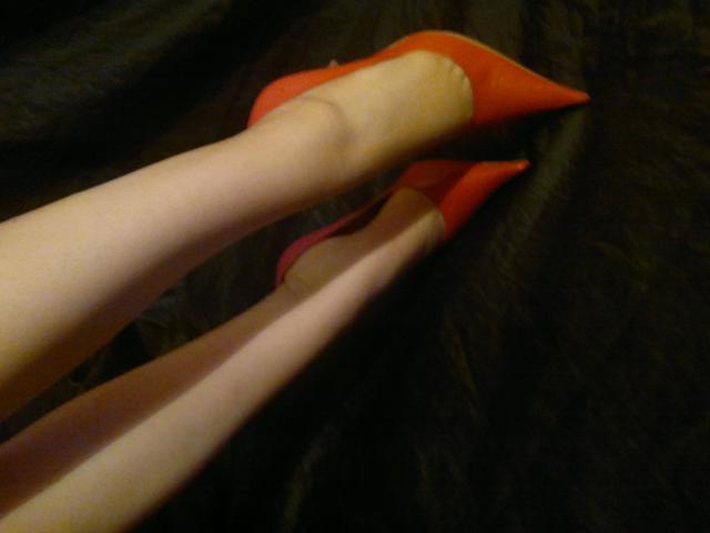 KatarinaArsch-hot's legs in sheer pantyhose with bright orange pointed heels, her feet crossed at the ankles on a dark, textured surface.