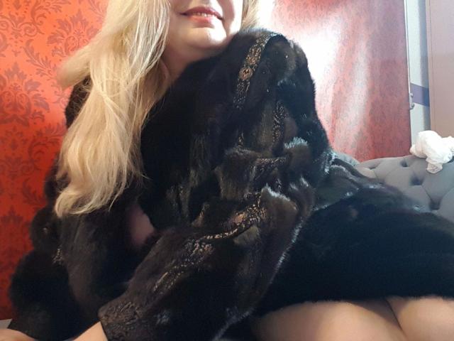 UntreueEhefrau41-hot with long blonde hair, wearing a black lace fur robe, sitting on a grey tufted couch, showing her ass, smiling with red lips.