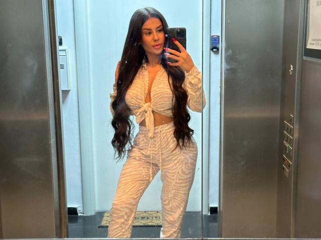 EvaZane, avec de longs cheveux bruns, portant un crop top blanc à rayures zébrées et un pantalon assorti, prend un selfie devant un miroir dans un ascenseur, montrant ses seins et ses fesses..