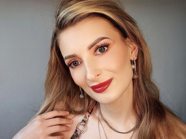 HeisseManuelle-hot ha i capelli biondi, il rossetto rosso e sta sorridendo con la mano vicino al viso. Indossa degli orecchini, una collana a catena e ha l'eyeliner arancione..