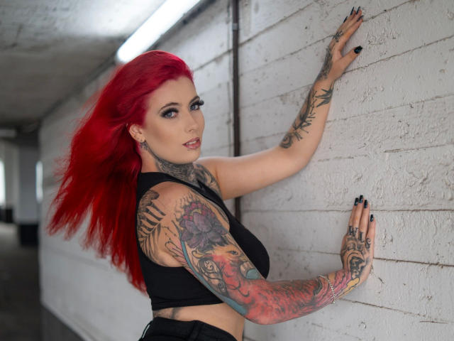 Bonnie-Stylez with bright red hair, tattoos covering arms and side, black crop top, black nails, hands on white brick wall, looking over shoulder, in dimly lit corridor.
