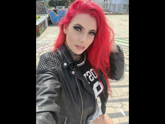 Bonnie-Stylez with bright red hair, wearing a black leather jacket with spiked shoulders, smiling at the camera. She's got tattoos on her neck and a sexy look.