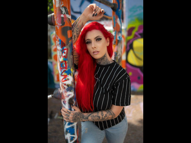 Bonnie-Stylez with bright red hair, tattoos, black pinstriped shirt, and blue jeans, leaning on a graffiti-covered pole, showing off her tattooed arms and confident, sexy look.