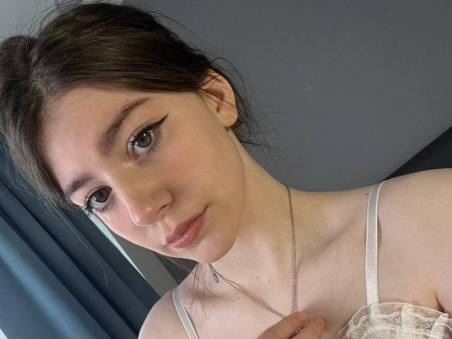 VinnaSkyHott mit heller Haut, braunen Augen und schwarzem Eyeliner, trägt eine weiße Spitzenbluse, hält eine silberne Halskette, hat ein leichtes Lächeln und braunes, zu einem unordentlichen Dutt gebundenes Haar..