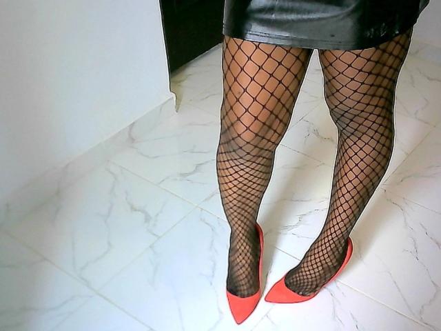 Goddessqueen-hot's legs in black fishnet stockings and red pointy-toe heels. She's wearing a black leather skirt, standing on white marble tiles. Her legs are sexy and hot.