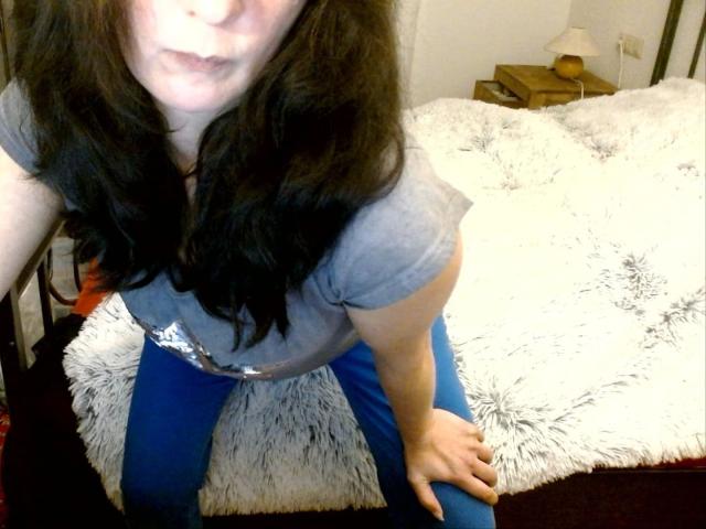 GeilesLuder-hot with wavy black hair, gray t-shirt, and blue pants, leaning forward on a furry white bed, showing a hand on her ass, slightly pouting lips.