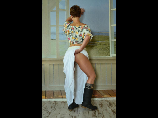 Bianca-Allein with short brown hair, floral crop top, white skirt, thigh-high slit, black boots, standing against a wall, looking over shoulder, showing her ass and toned legs.