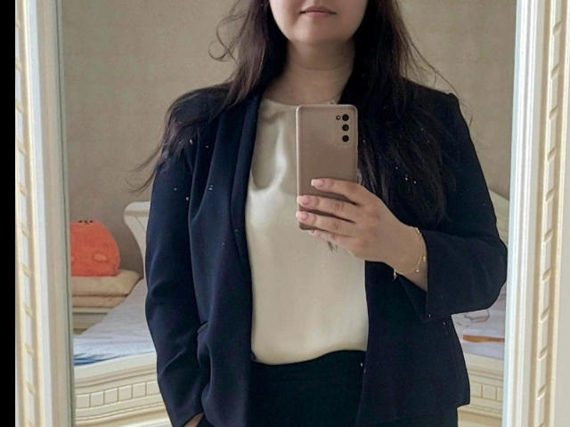 Goldmoon-hot with long dark hair, wearing a black blazer over a white top, holding a gold iPhone, standing in front of a mirror, hands in pockets.