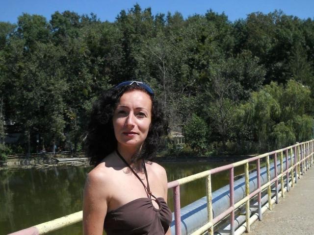 Pussyxxx-hot with curly black hair, wearing a brown halter top, standing on a bridge with a metal railing, and blue sunglasses on her head, smiling at the camera.