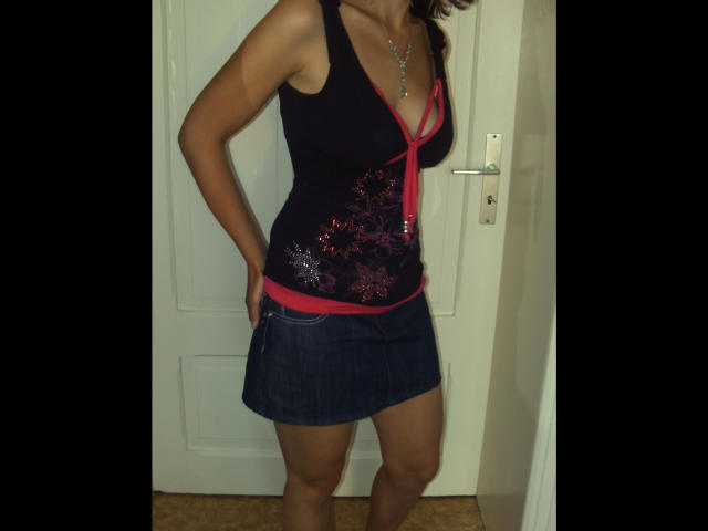 Fickmaus12-hot with tan skin, black tank top, red trim, sparkly design, blue denim skirt, hands on hips, necklace, visible breasts, standing in front of white door.