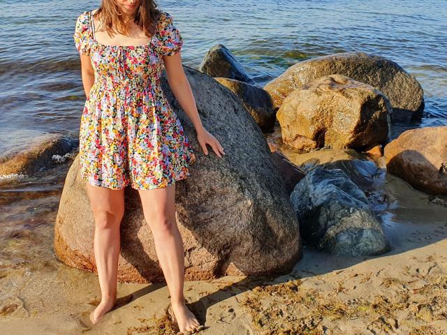 Fickmaus12-hot with floral dress, standing barefoot on sandy beach, leaning against large rock, water in background, rocks in foreground, sunlight casting shadows, brown hair, floral pattern.