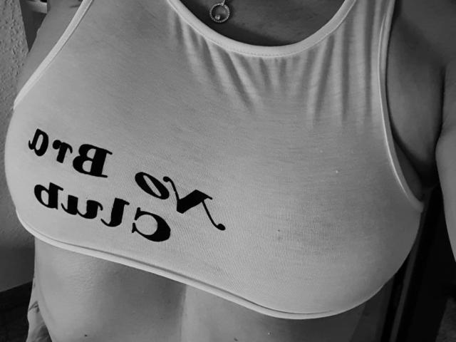 AbenteuerMelody-hot's large, round tits are visible in a gray sports bra with black text. She wears a small necklace and the bra is slightly lifted. Black-and-white photo.