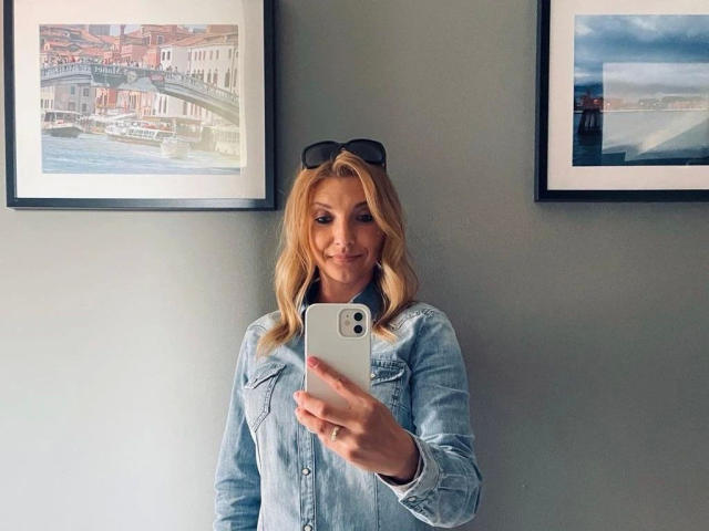 AbenteuerMelody-hot with blonde hair, wearing a blue denim shirt, holding a white iPhone, smiling, black sunglasses on her head, standing in front of two framed pictures.