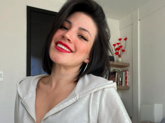 Liliysweet has medium tits, black shoulder-length hair, red lipstick, white hoodie, and silver hoop earrings, smiling at the camera with her head slightly tilted.