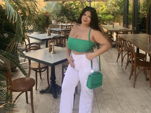 JulietaStark with green strap top, white pants, green crossbody bag, long dark hair, red lipstick, medium tits, hand on hip, standing in outdoor cafe with wooden chairs.