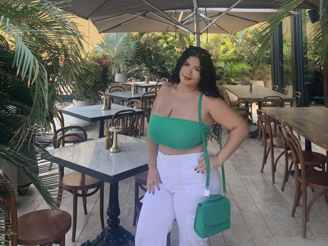 JulietaStark with curly black hair, green crop top showing her big tits, white pants, green purse, standing by outdoor tables, surrounded by plants and wooden chairs.