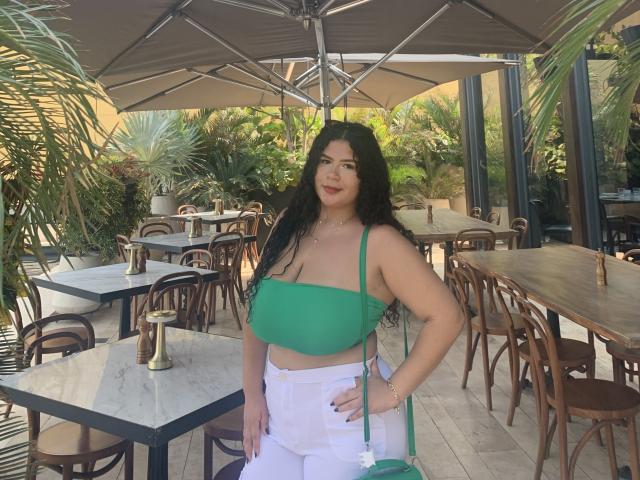 JulietaStark has long black hair, green tube top showing her boobs, white pants, and a green shoulder bag, standing outdoors at a cafe with wooden chairs and tables.