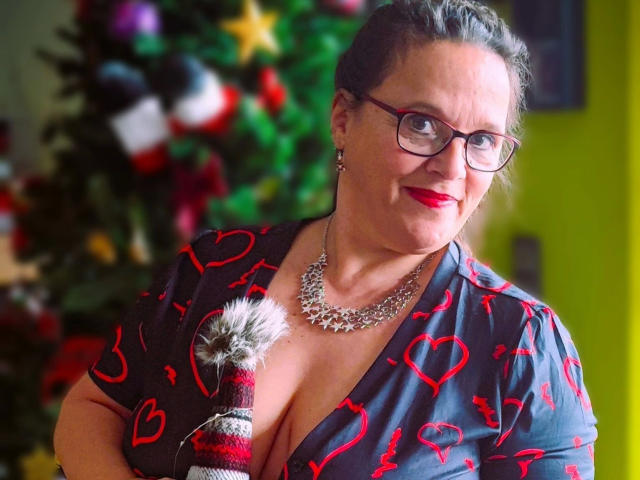 Lady-Mercedes with glasses, red lipstick, and a necklace, wearing a grey shirt with red heart patterns, holding a fuzzy red and white pom-pom, showing her big boobs.