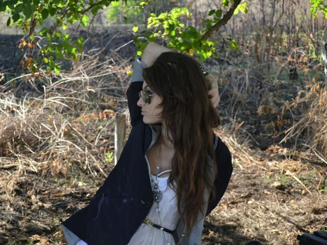 Lovly19-hot with wavy brown hair, wearing a dark jacket over a white dress, raising arms under green leaves, wearing sunglasses, standing in dry, grassy area.
