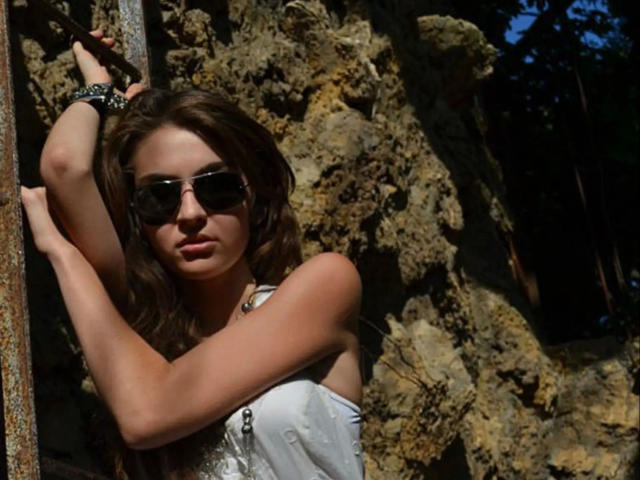 Lovly19-hot with wavy brown hair, wearing sunglasses and a white top, leans against a rusty metal structure, showing her tits and a black wrist bracelet in sunlight.