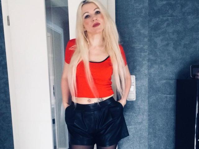 Sexicandy-hot with long blonde hair, red crop top, black leather shorts, hands in pockets, tattoo on belly, looking up with red lipstick, standing against gray wall.