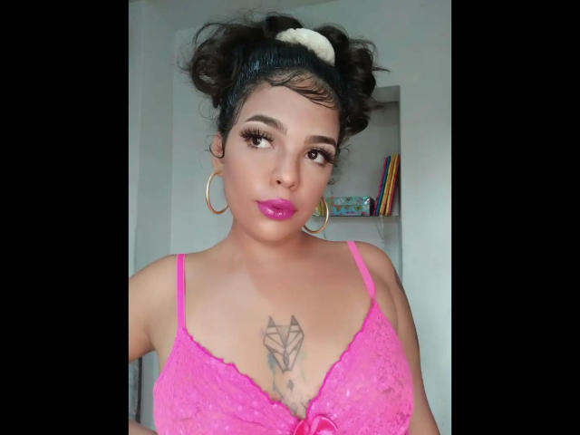 Valkirie-hot with dark curly hair, pink lipstick, hoop earrings, and pink lace bra showing her tits and a diamond tattoo above her boobs. She looks cute and confident.