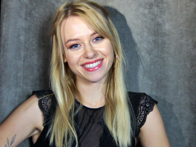 Cindy-Lust, blonde with straight hair, smiling with pink lipstick, black lace sleeve top, blue eyes, small tattoo on left upper arm, against gray background.