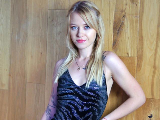 Cindy-Lust has blonde hair, blue eyes, and pink lipstick. She's wearing a sparkly black dress, showing tattoos on her left arm, and a necklace with a small pendant.
