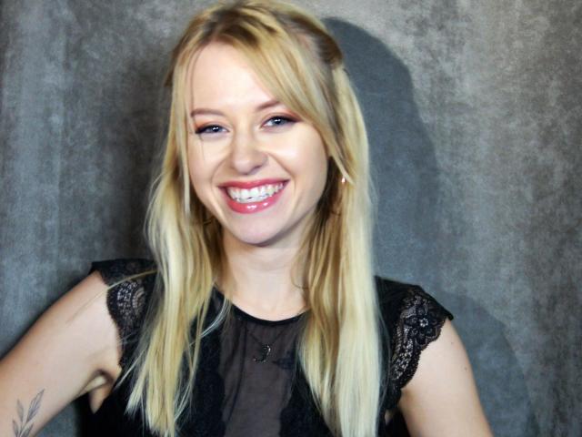 Cindy-Lust has blonde hair, blue eyes, and a cute smile. She's wearing a black lace top, showing a small tattoo on her right arm. Her tits are small.