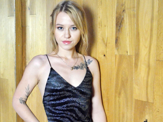Cindy-Lust with blonde hair, blue eyes, and pink lipstick, wearing a black, sparkly, V-neck dress, showing tattoos on her arm and chest, standing against wooden panels.