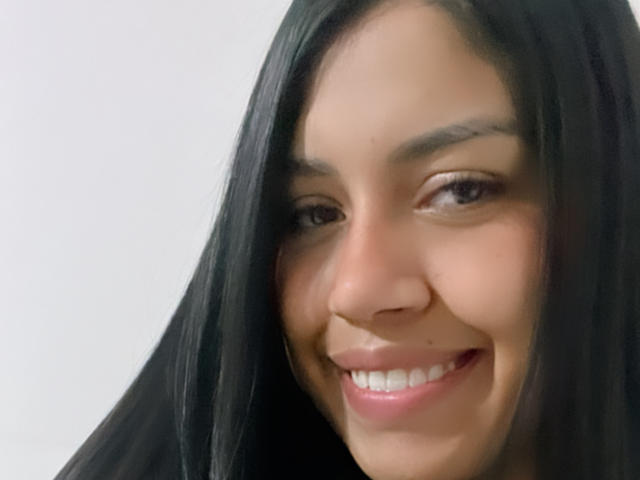 Stephanie Davies tiene una piel marrón lisa, cabello negro largo y una sonrisa encantadora con dientes blancos. Sus ojos marrones están ligeramente entrecerrados, mostrando una expresión relajada y feliz..