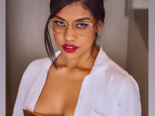 StephanieDavies with brown skin, dark hair, glasses, red lipstick, white shirt, and visible cleavage, looking directly at the camera.