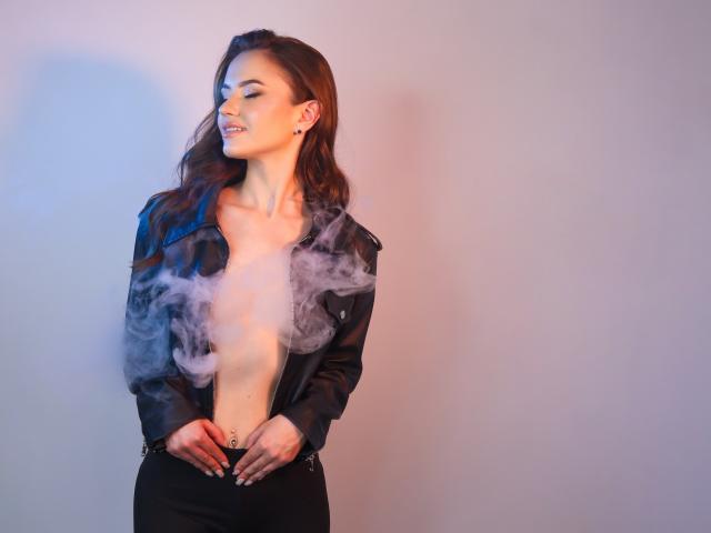KaraCriegs with smoky breath, eyes closed, black leather jacket open, showing tits, black pants, wavy brown hair, earrings, standing, soft blue and pink light.